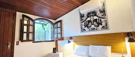 Un dormitorio con techo de madera, cama con ropa de cama blanca, mesita de noche con lámpara y ventana con vistas a la vegetación.