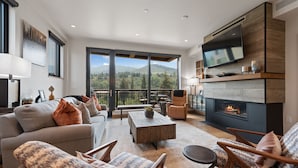 A modern living room with a fireplace, comfortable seating, and a large window overlooking a scenic mountain view. - Riverfront Lodge (Avon)