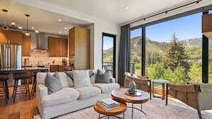 A modern living room with a sofa, coffee table, and chairs, a kitchen with wooden cabinets, and a large window offering a mountain view. - Riverfront Lodge (Avon)