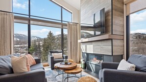 A modern living room with a fireplace, large windows, and a view of mountains. - Riverfront Lodge (Avon)