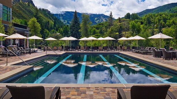 A pool area with lounge chairs and umbrellas, surrounded by lush greenery and mountains in the background. - Riverfront Lodge (Avon)