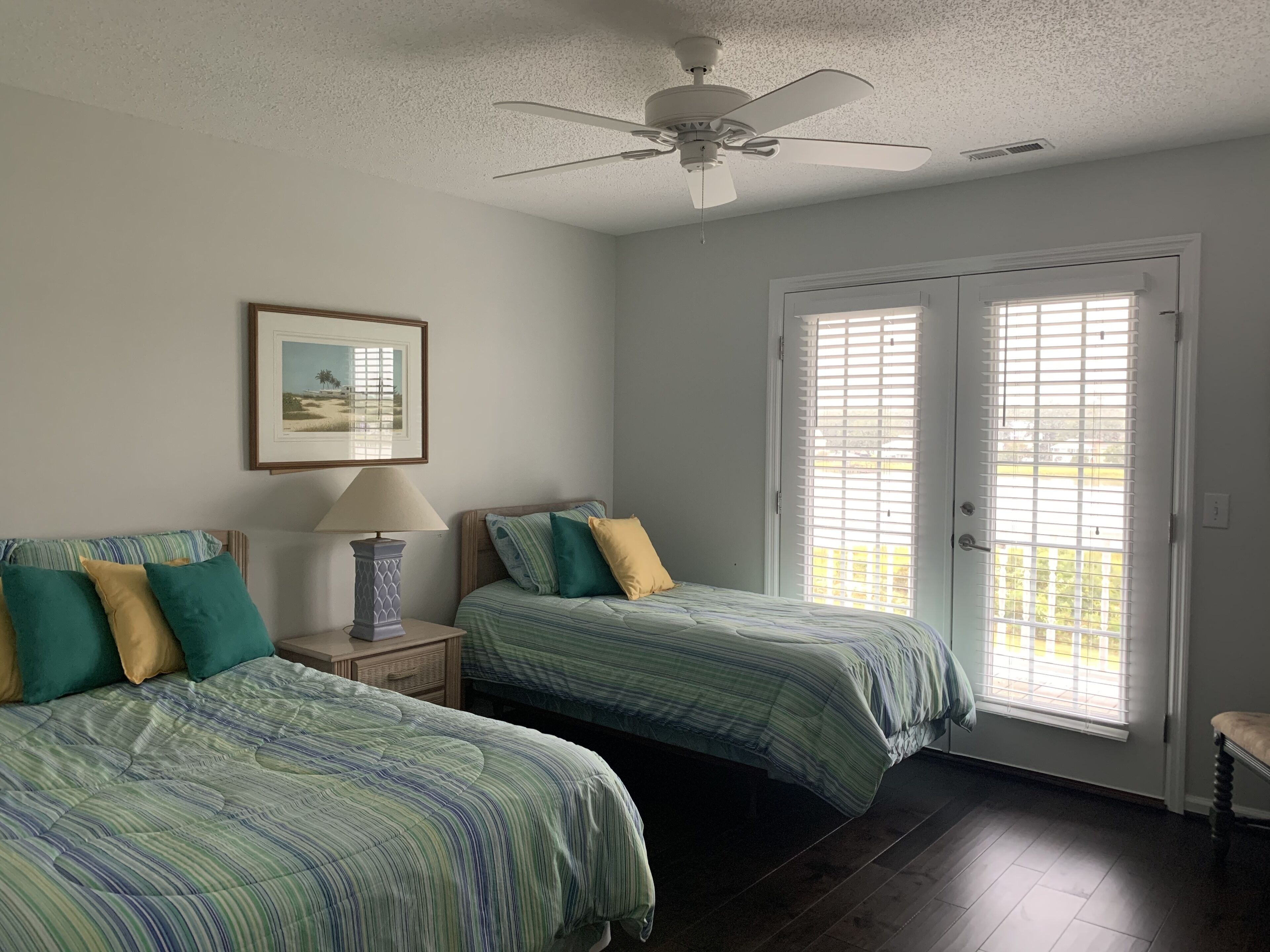 A bedroom with two beds, a ceiling fan, a framed picture on the wall, and a door with blinds.