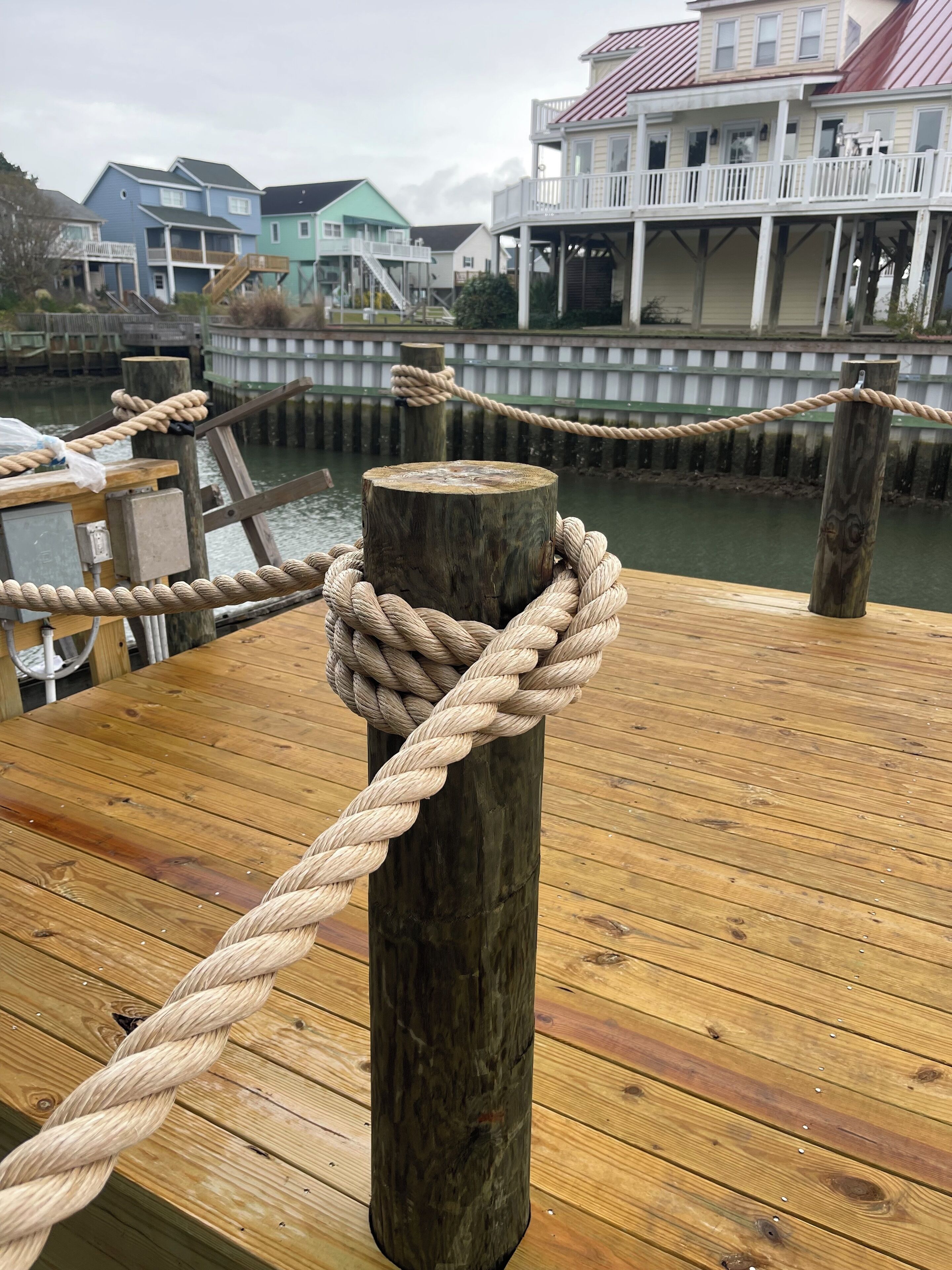 A wooden dock with a rope tied around a post, wooden planks, and a view of houses in the background.