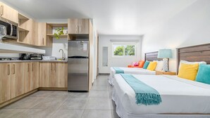 Een moderne hotelkamer met kitchenette, twee bedden met kleurrijk beddengoed, een nachtkastje en een raam met uitzicht op groen.