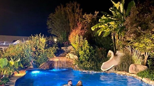 En swimmingpool med kristallklart vatten, omgiven av tropiska växter och en dekorativ vattenfallsanläggning.