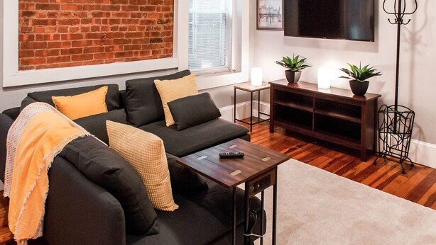 A living room with a black sectional sofa, a wooden coffee table, a TV mounted on the wall, and a brick wall. - Gorgeous Wooster Sq. apt. - wshr/dryr, parking (New Haven)