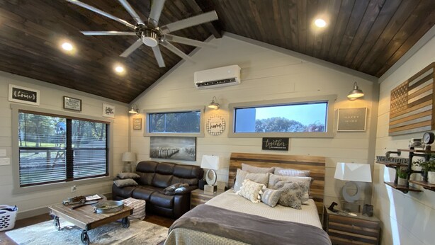 A spacious bedroom with a large bed, a leather sofa, and a desk. There is a ceiling fan, an air conditioning unit, and a window with blinds. - Circle C Cabin Perfect for Cozy Relaxing Retreats (Caldwell)