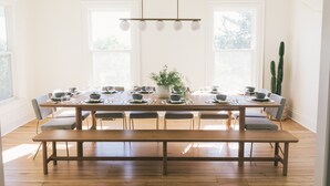 Comedor con una larga mesa de madera dispuesta para seis comensales, rodeada de sillas. Hay un banco en el centro y una planta en maceta sobre la mesa.