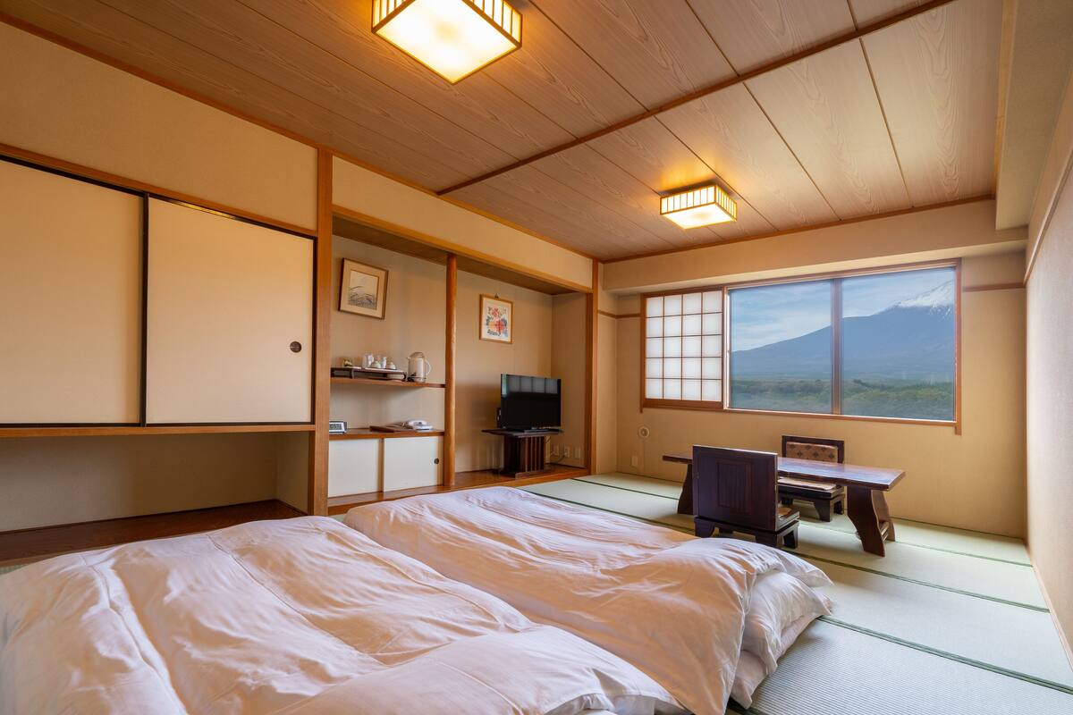 Una tradizionale stanza giapponese con pavimento in tatami, un letto, una TV, una scrivania e una finestra con vista su una montagna.