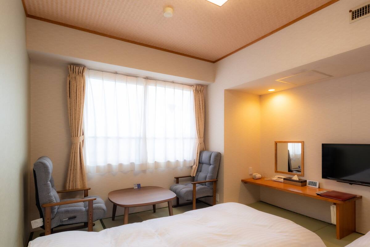 Camera d'albergo con due letti, una scrivania, una sedia, un tavolino, una televisione e una finestra con tende.
