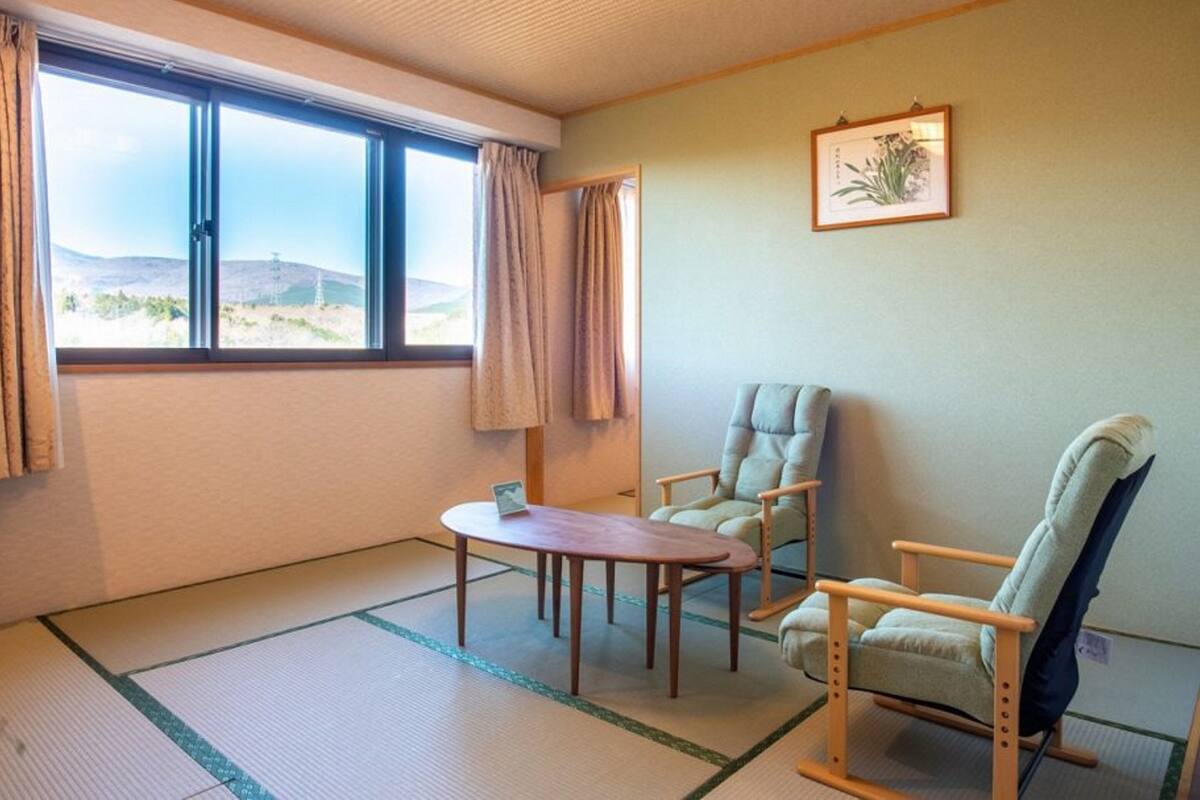 Una stanza con tatami, un tavolo, due sedie, una finestra con vista sulle colline e un quadro appeso al muro.