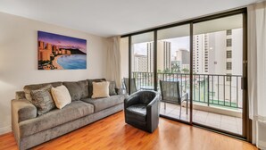 A modern living room with a grey sofa, a black leather chair, and a large window with a cityscape view. - Block to Beach! Recreation Deck View Deluxe Condo, Free parking & Wifi (Honolulu)
