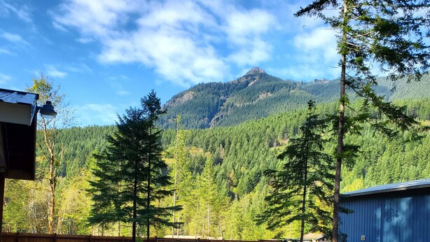 A gravel parking area with a wooden fence and a blue building, surrounded by tall evergreen trees and a mountain backdrop. - RV Flat Lot- Tatoosh Timbers - No Bedroom or Bathroom RV Lot only.  (Packwood)