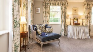 A room with a dark blue sofa, floral curtains, a small table with a lamp, and a window with a view of greenery. - The Courtyard House, Dorchester (Dorchester)