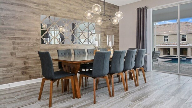 A dining area with a wooden table and chairs, a decorative wall, and a view of a swimming pool. - Ultimate New 8 Bedroom 5 Bathroom Champions Gate Resort Property with Spa and Pool (Davenport)