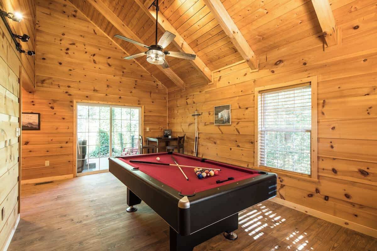 A wooden interior with a pool table, ceiling fan, and large windows.