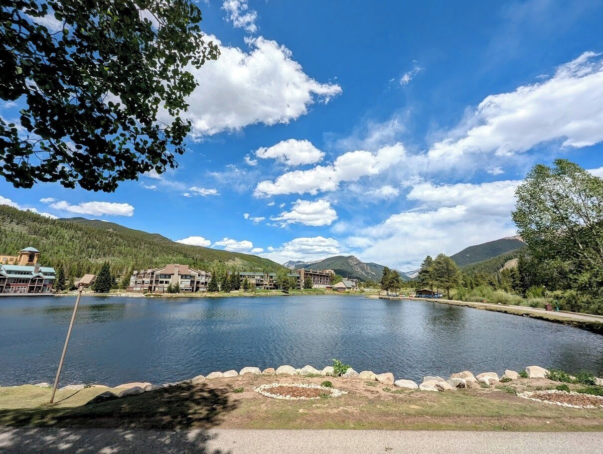 A lakeside view with buildings, a clear sky, and mountains in the background.