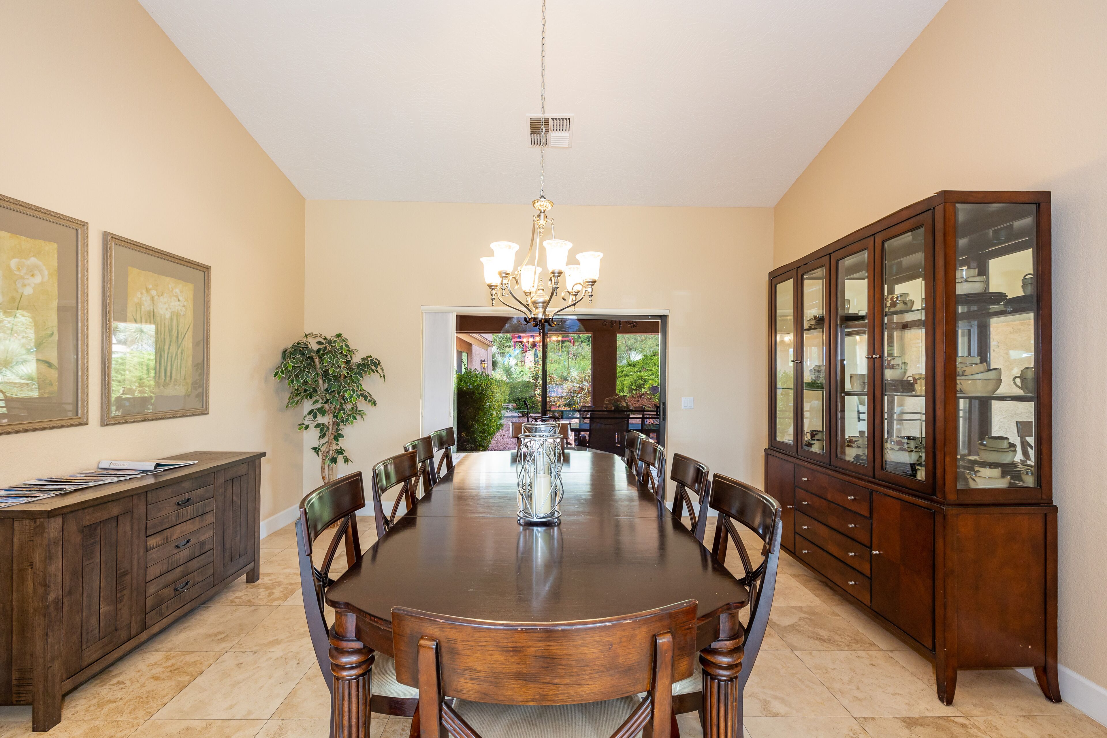 A dining room with a large wooden table, chairs, a chandelier, a cabinet with glass doors, and framed artwork on the walls.