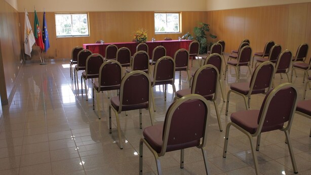 Un salón de conferencias con filas de sillas, una mesa con mantel rojo y banderas en la pared.