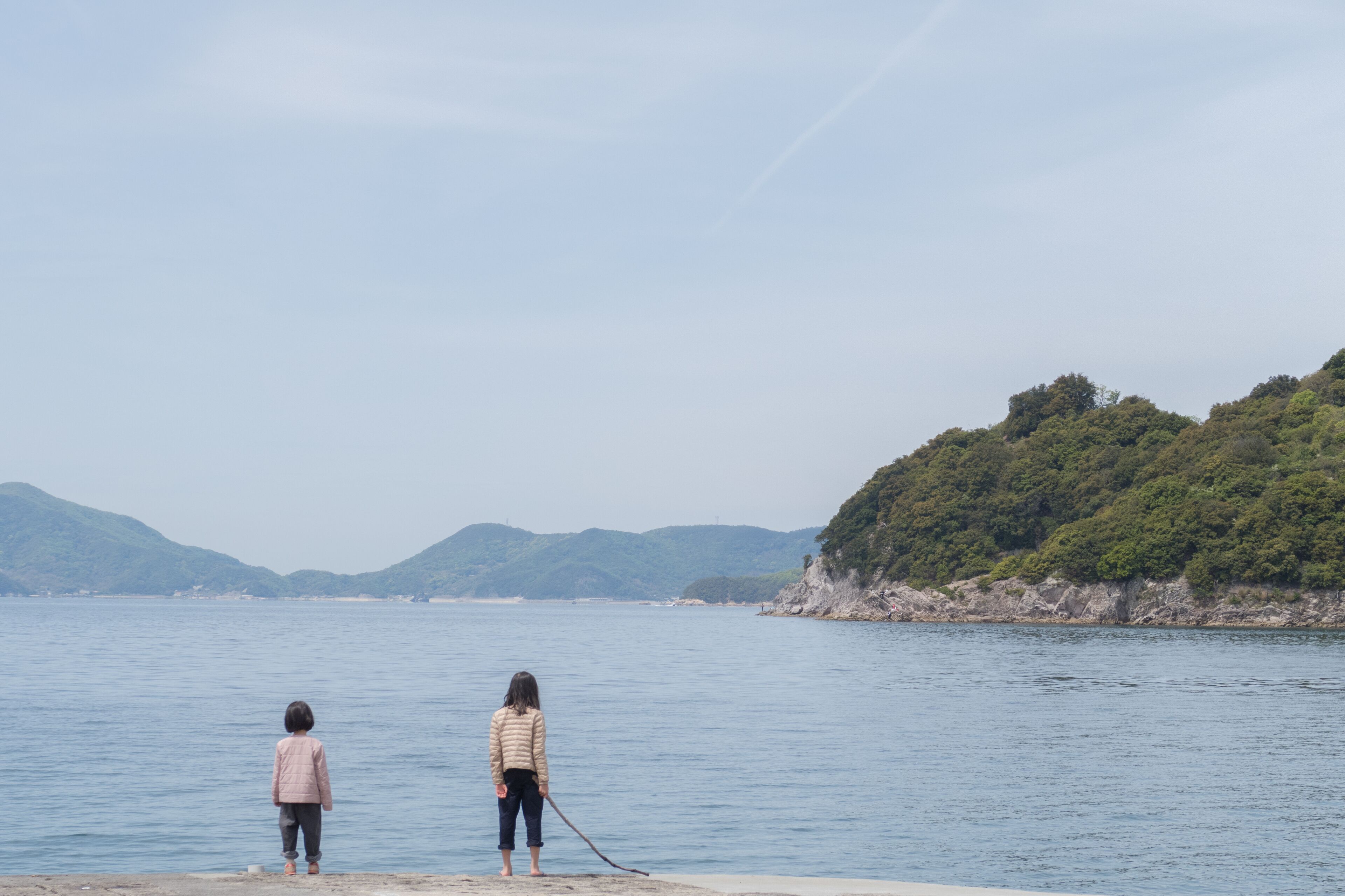 Dos personas de pie junto a un cuerpo de agua tranquilo con un paisaje montañoso al fondo.
