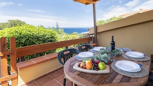 Eine Holzterrasse mit gedecktem Tisch, mit Blick auf eine malerische Landschaft aus Meer und Grün.