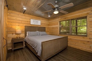 Une chambre à coucher aux murs lambrissés, comprenant un lit, des tables de chevet, un ventilateur de plafond et une fenêtre donnant sur un paysage agréable.