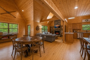L’intérieur d’une cabane en rondins comprend un coin repas, un salon, une cheminée, une cuisine et de grandes fenêtres.