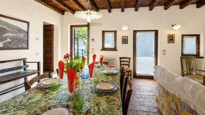 Uma área de refeições com uma mesa posta para quatro pessoas, vigas de madeira no teto e vista para o exterior através de uma janela.