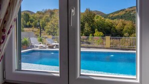 Vista a través de una ventana que muestra una piscina con sillas de descanso y un paisaje montañoso.