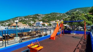 Un parque infantil con tobogán y columpios con vistas a un puerto deportivo con barcos y un pueblo en la ladera de una colina al fondo.