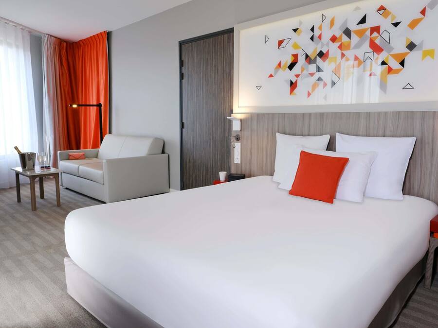 A hotel room with a large bed, a sofa, a nightstand, and a wall-mounted lamp.
