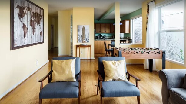 Un salon avec deux fauteuils bleus, une table basse en bois et une table de baby-foot. Au fond, on aperçoit une cuisine avec des meubles en bois et une fenêtre.
