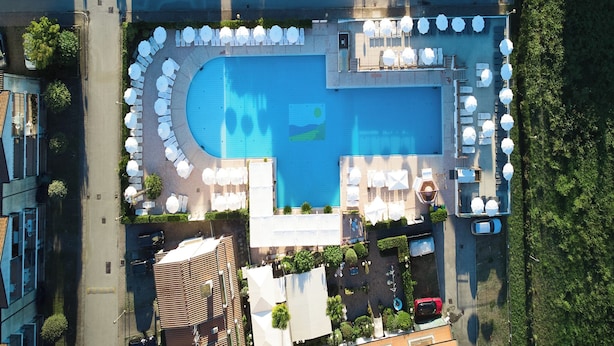 An aerial view of a hotel with a central swimming pool surrounded by sun loungers and umbrellas, a parking area, and a small garden area with a water feature. - Cozy Flat in a Bright Residence-beahost (Rosolina)