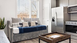 A modern living room with a grey sofa, a wooden coffee table, and a kitchen area with a stainless steel refrigerator and oven.