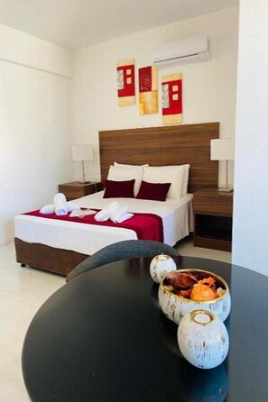 Quarto de hotel com uma cama bem arrumada, uma mesa redonda preta com vasos decorativos e uma cabeceira de madeira.