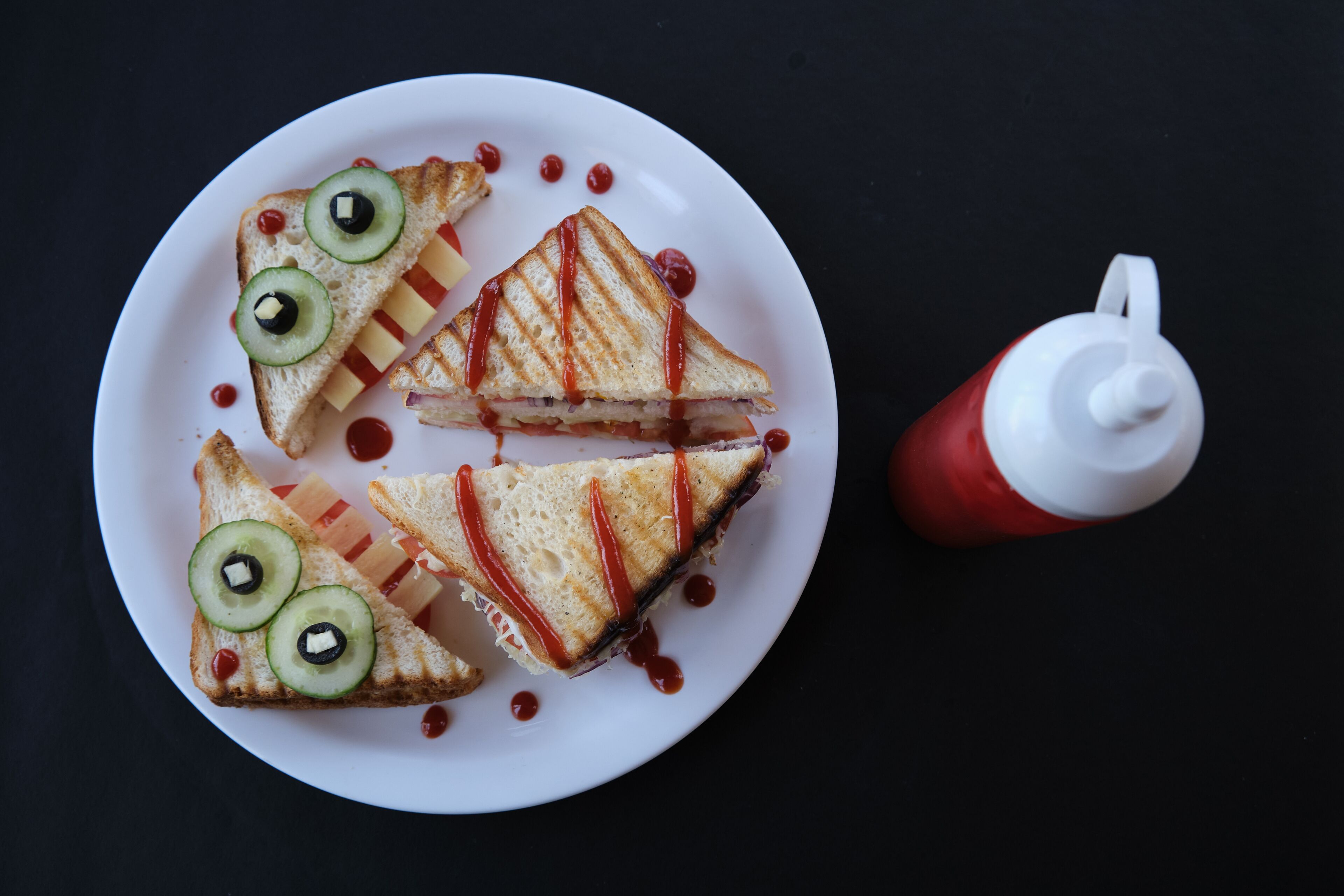 Un plato con un sándwich a la plancha, rodajas de pepino, kétchup y una botella de kétchup.