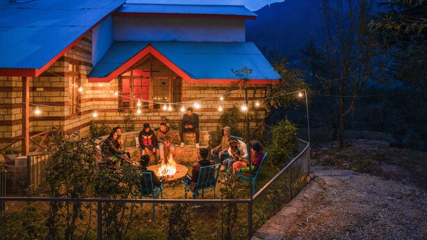 Un chalet rustique aux murs en pierre et au toit rouge, entouré d’un jardin et d’un foyer où des personnes sont rassemblées.