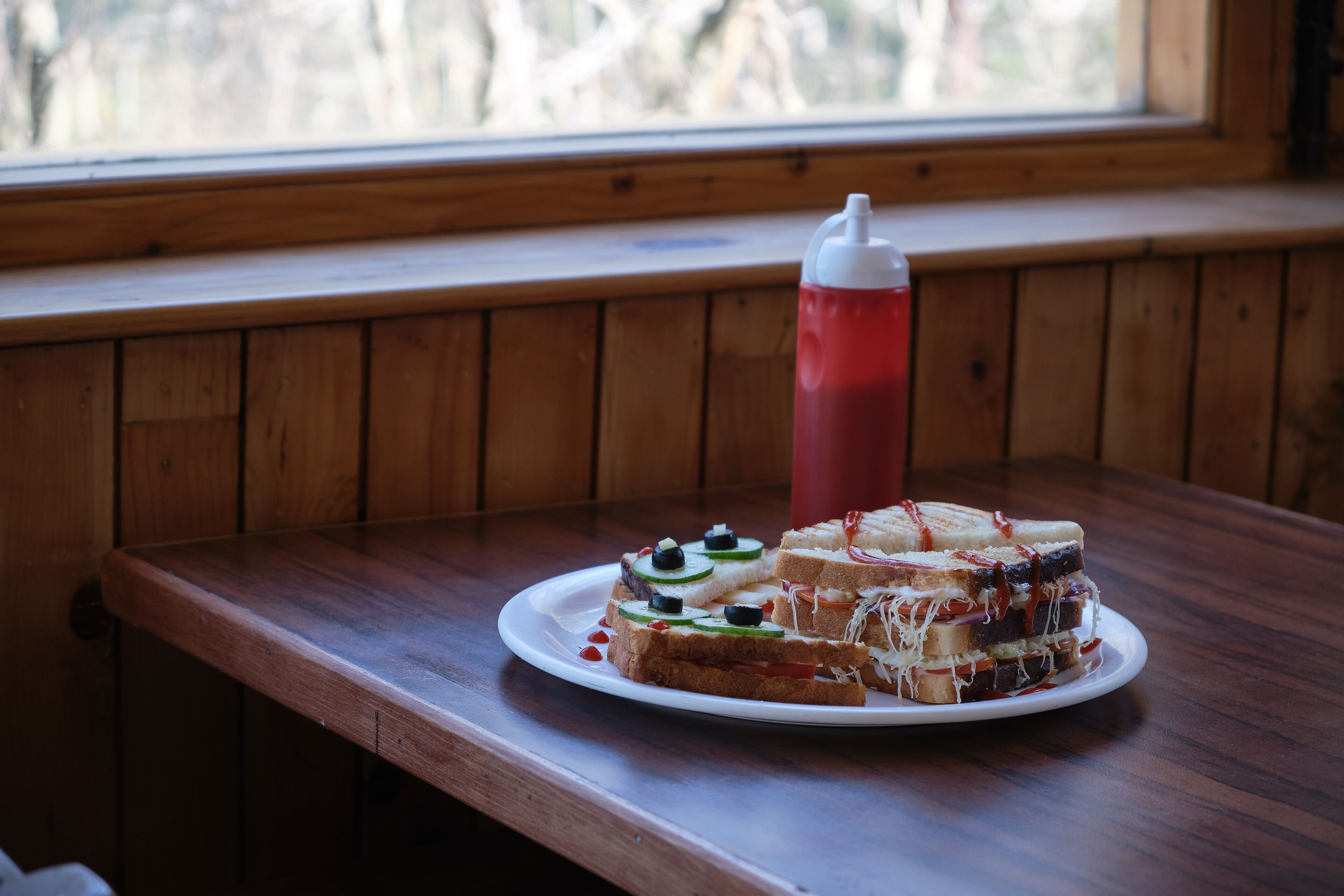 Un sandwich sur une assiette, avec une boisson rouge sur la table, près d’une fenêtre.