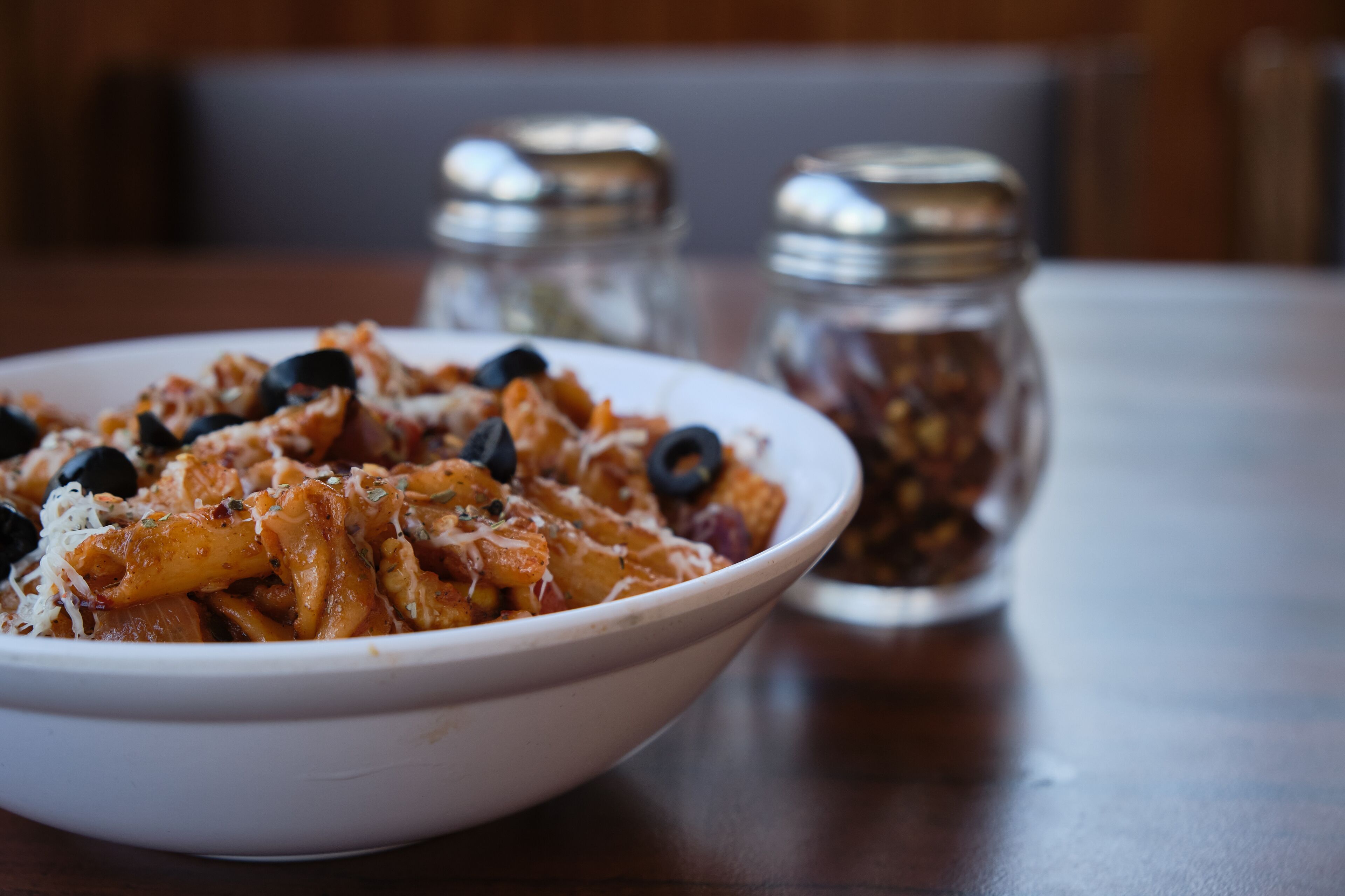 A bowl of pasta with black olives and cheese on a table.