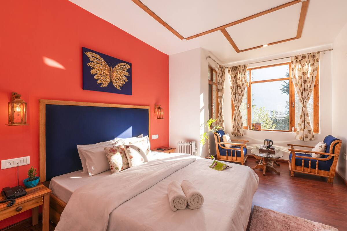 A hotel room with a large bed, a wooden headboard, a window with curtains, a small table, two chairs, and a wall art piece.