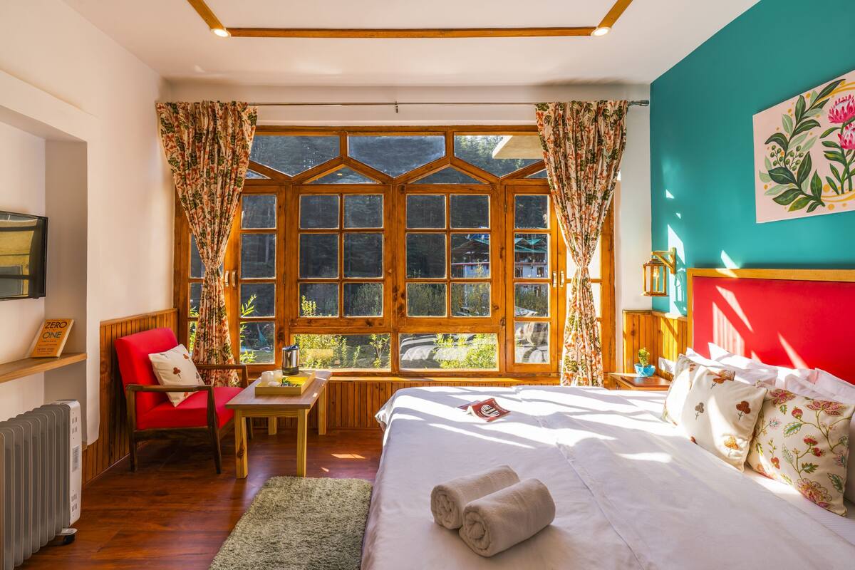 A room with a large window, a bed, a red chair, a table, and a radiator.