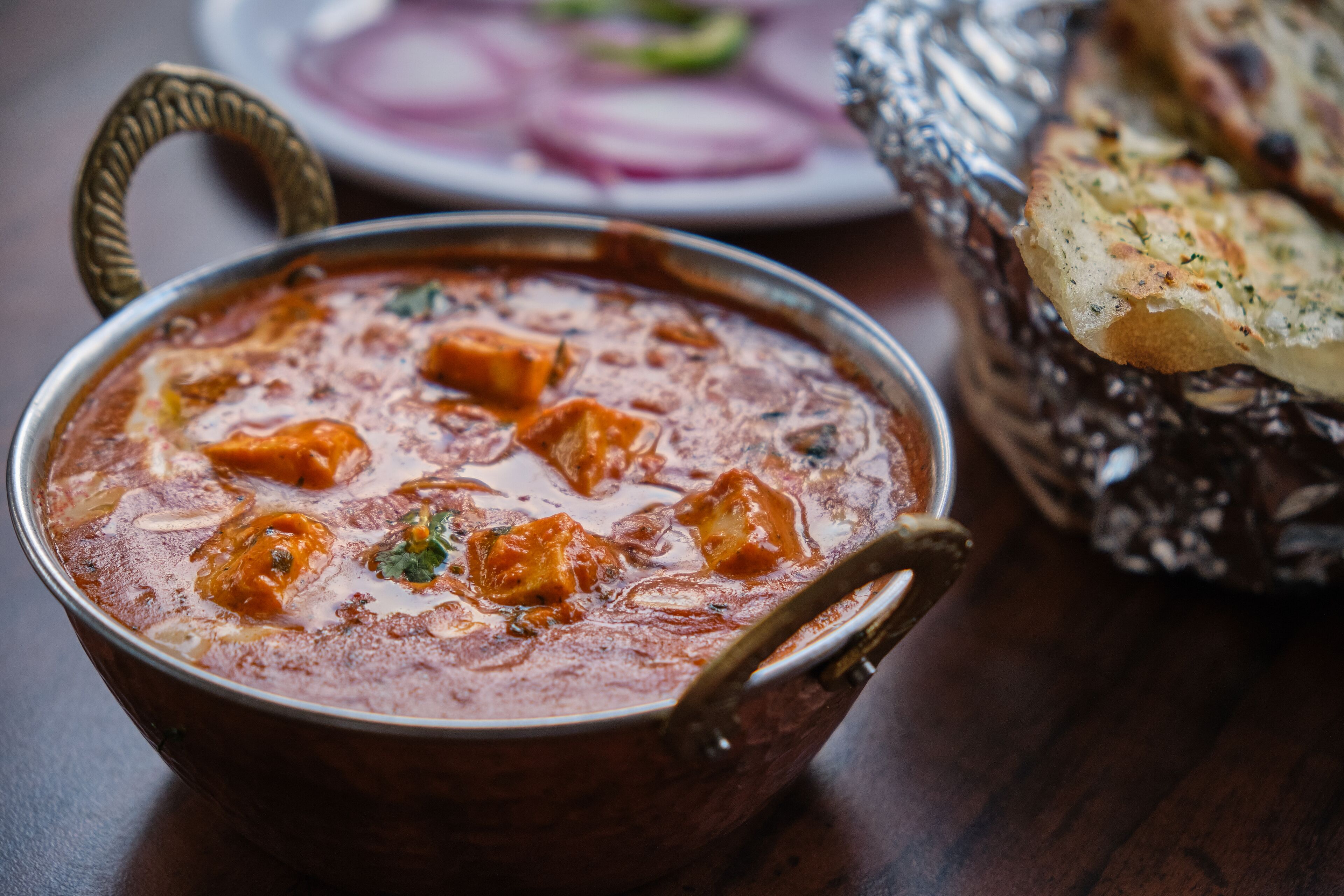 A bowl of curry with a spoon, accompanied by a plate of sliced onions and a piece of bread.