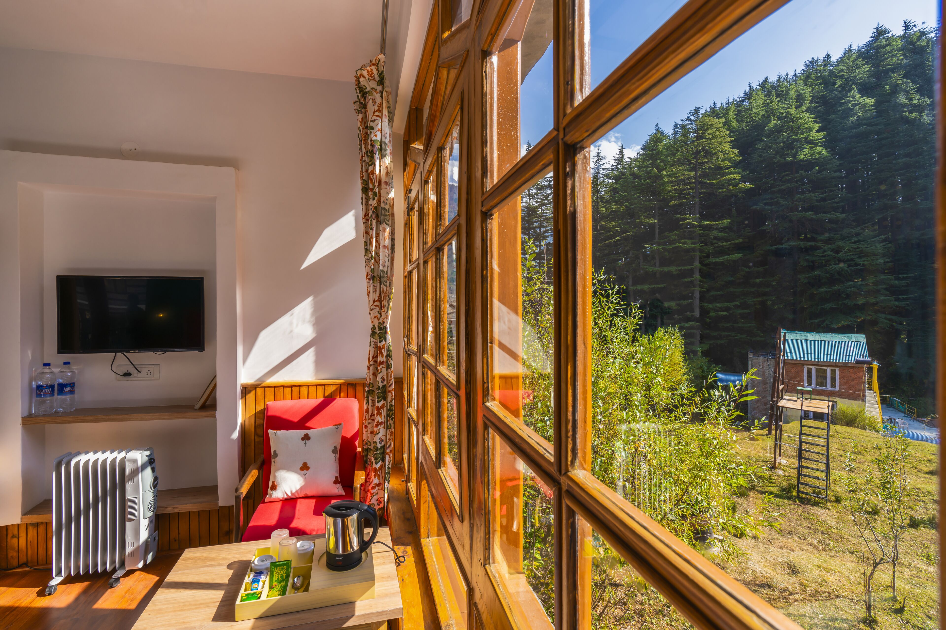 A room with a bed, a radiator, a table with a kettle, glasses, and a tray of food. There is a TV on the wall and a window with a view of a forest and a small building outside.