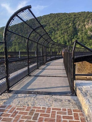 A pedestrian bridge with a metal railing and brick pathway, leading to a forested hillside.