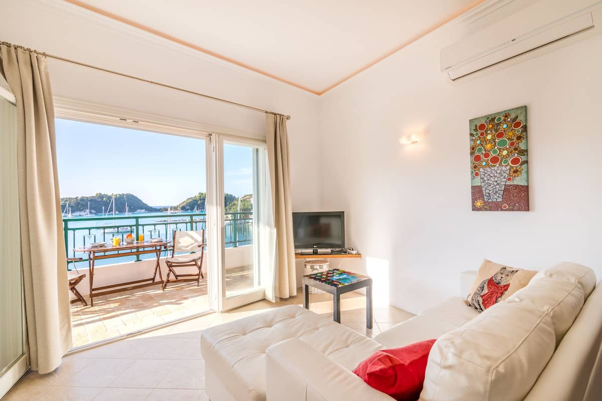 Un salon avec un canapé, une table basse et un téléviseur. Il y a un balcon avec vue sur la mer et les montagnes.