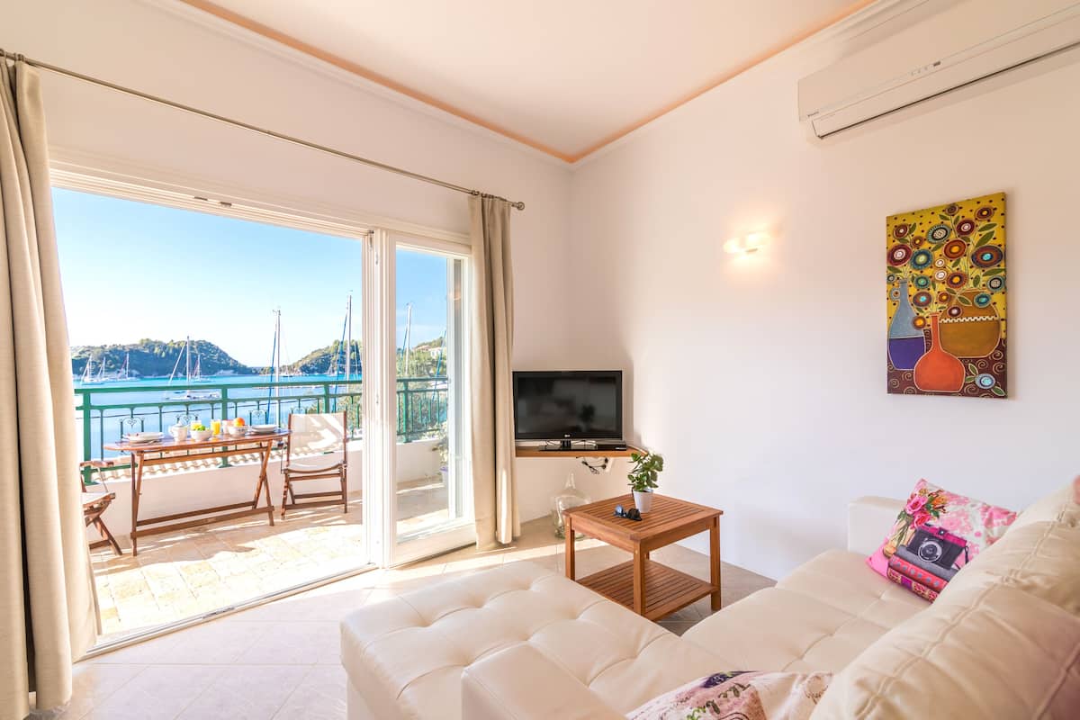 Un salon comprenant un canapé, un téléviseur, une petite table avec une plante, et un balcon avec une table et des chaises.