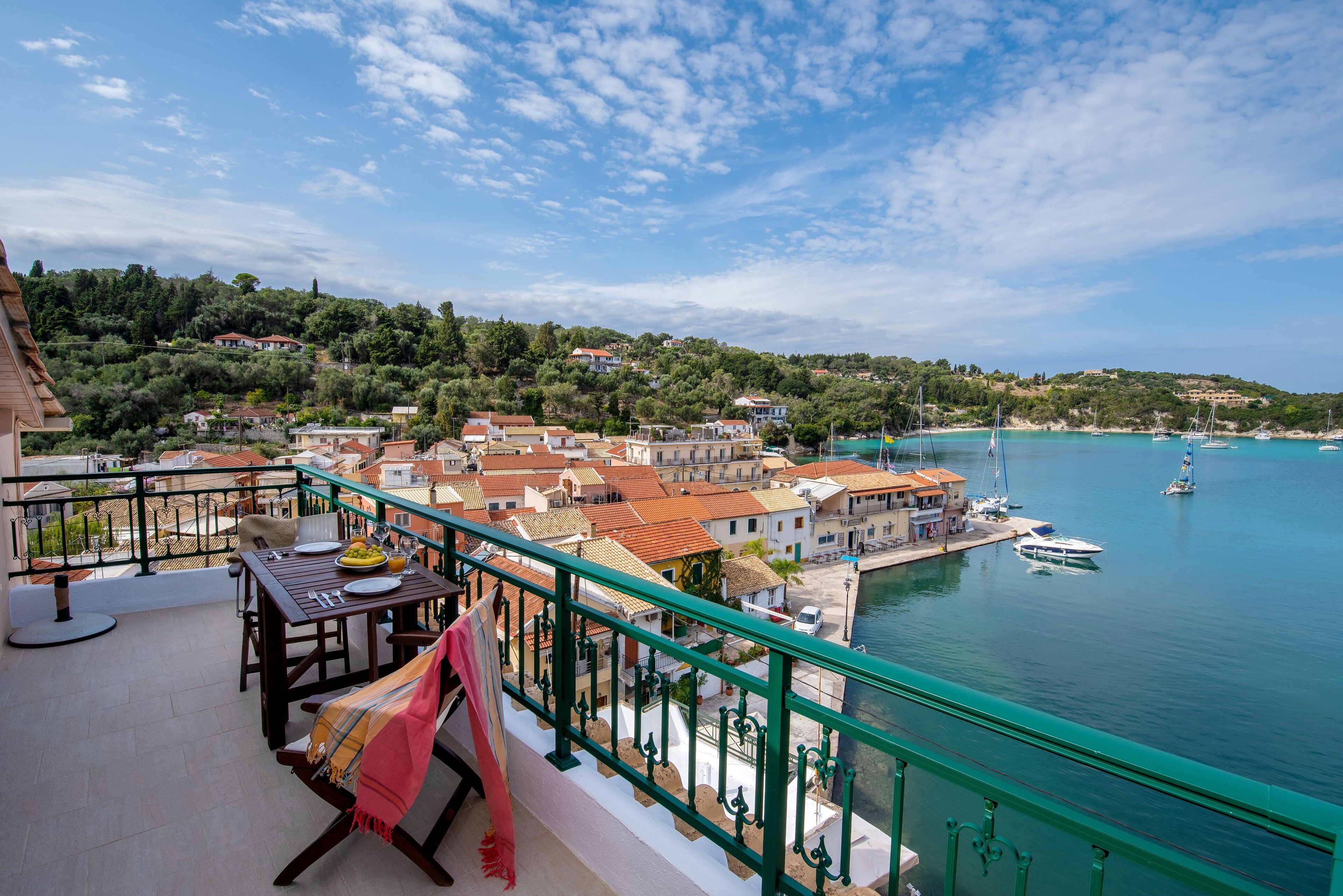 A balcony with a table set for dining, overlooking a coastal town with buildings and boats.