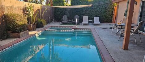 A rectangular swimming pool with a tiled edge, surrounded by a concrete deck. There are lounge chairs and a table with a green umbrella.