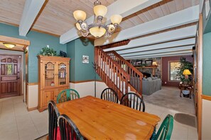 A dining area with a wooden table and chairs, a chandelier, and a staircase leading to a living room. - Peace Out by Destination Big Bear : Remarkable Dutch Gambrel Only 2 Blocks from Snow Summit! Foosball! (Big Bear Lake)