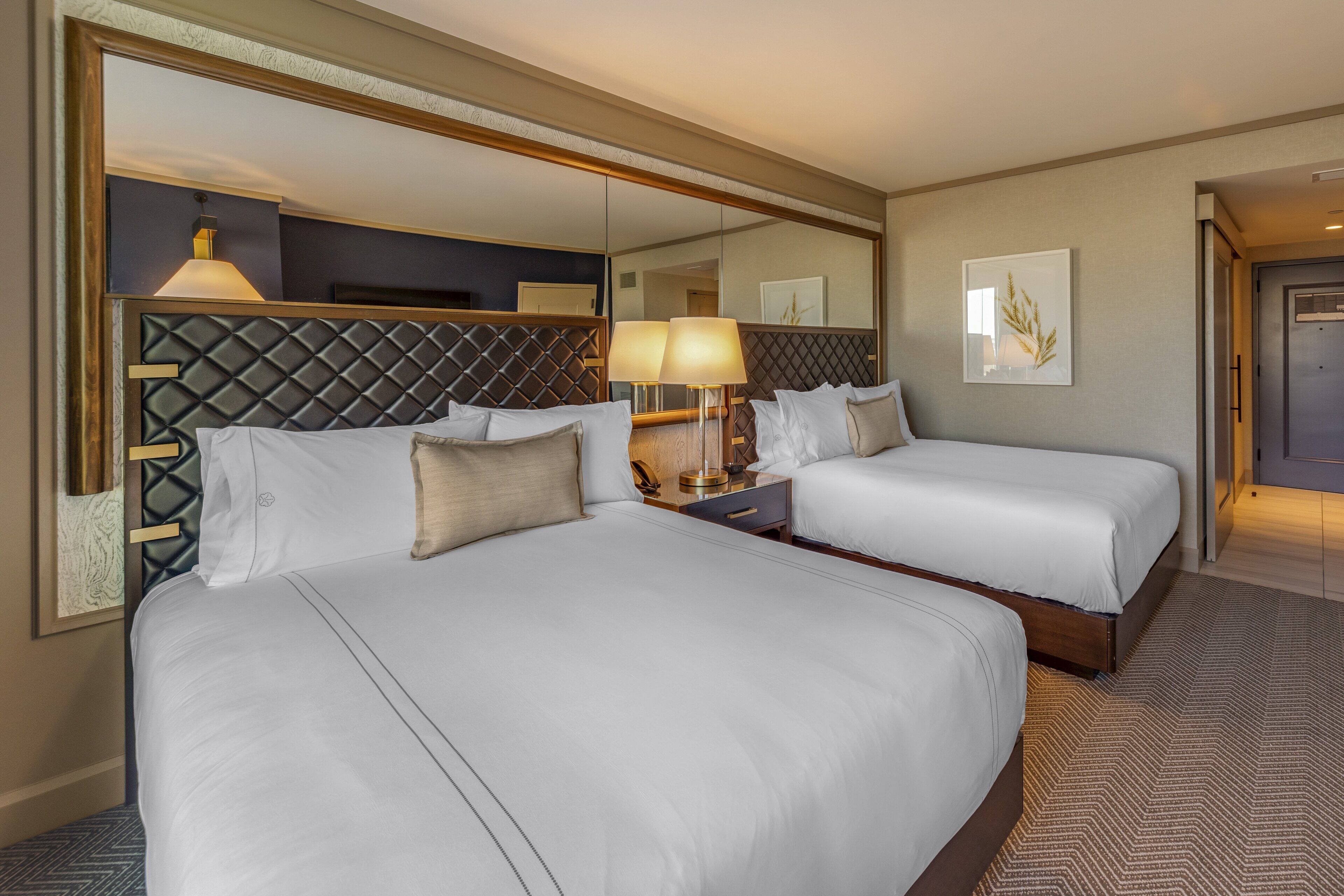 A hotel room with two beds, a large mirror, a bedside table with a lamp, and a framed picture on the wall.
